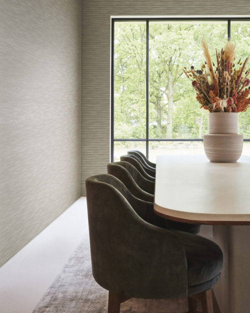 Une salle à manger sereine baignée dans des tons de beige et de gris clair. À droite, une table élégante entourée de chaises confortables. La table est ornée d'un vase en céramique contenant un bouquet de fleurs séchées. Un tapis gris délimite l'espace et mène à une grande fenêtre au fond, qui offre une vue pittoresque sur les arbres. Les murs sont revêtus de la collection Rivage haute performance d'Omexco en gris clair, qui présente une structure subtile et délicatement surélevée évoquant les contours tranquilles d'un horizon lointain. Ces revêtements muraux sans PVC offrent non seulement la possibilité d'être nettoyés à l'eau de Javel, mais aussi des performances accrues en matière de résistance au frottement, garantissant à la fois style et durabilité.