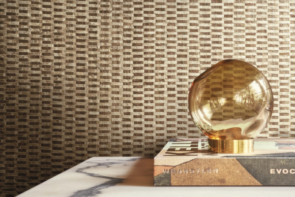 Une élégante lampe de table ronde en verre, joliment placée au-dessus d'un livre, sert de point focal à ce décor élégant. La toile de fond est constituée de luxueux revêtements muraux en capiz de la collection Archipelago d'Omexco, qui présentent un mélange captivant de tons dorés et bruns, créant ainsi une atmosphère sophistiquée et accueillante.