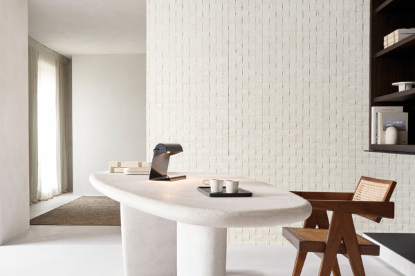 Un espace bureau sophistiqué mettant en avant la collection Pure d’Omexco, référence PU281, un revêtement mural audacieux qui présente un tissage d’écorce de mûrier, mettant en valeur la richesse et la texture inhérentes de ce matériau naturel. Le design en grille carrée complexe ajoute un élément tactile et une profondeur visuelle à l’espace, soulignant la beauté organique de l’écorce. La pièce comprend un bureau sculptural blanc aux courbes douces, orné d’une lampe de bureau noire moderne et d’un plateau contenant des tasses en céramique minimalistes. À droite, une chaise en bois inspirée du style des années 50 avec des détails en cannage complète les tons naturels du papier peint. Des étagères intégrées présentant des livres et des objets décoratifs s’intègrent harmonieusement à l’esthétique minimaliste globale, créant un espace de travail paisible et élégant.
