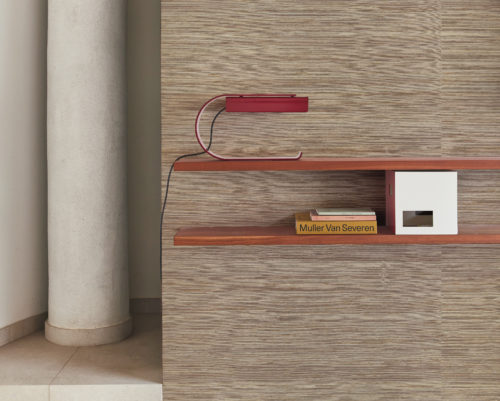 Deux étagères murales en bois ornées d'un livre de Muller van Severen et d'une lampe de table rouge élégante constituent un arrangement décoratif élégant. À gauche, une colonne beige ajoute une touche de simplicité moderne. En arrière-plan, la collection de revêtements muraux naturels Intuition d'Omexco, fabriqués à la main, se caractérise par un tissage en ramie dans des tons marron clair, créant une atmosphère chaleureuse et accueillante.