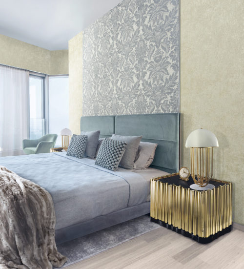 Chambre à coucher bleu clair avec des tableaux texturés et un plaid en fourrure. Table de nuit dorée avec lampe dorée. Au mur, un revêtement mural damassé bleu clair d'Omexco.