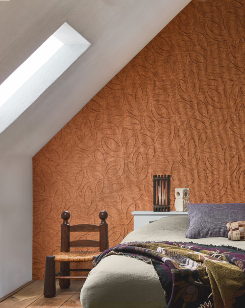 Cette chambre mansardée et chaleureuse met en valeur la référence OAS223, intitulée Verano, issue de la collection Oase d’Omexco. Ce revêtement mural est réalisé en tissage de papier raffiné, brodé d’un subtil motif organique qui accentue son élégance naturelle. Les tons chauds et terreux de l’orange s’harmonisent parfaitement avec le parquet rustique et le mobilier artisanal. La literie en lin doux, agrémentée de plaids et de coussins texturés dans des nuances profondes et feutrées, crée une atmosphère à la fois apaisante et accueillante. Une figurine sculpturale de hibou et une lampe lanterne ajoutent une touche artisanale, conférant à cet espace une ambiance intime et raffinée.