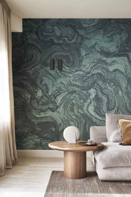 Un salon décoré avec une esthétique moderne. À gauche, une grande fenêtre encadrée de rideaux permet à la lumière naturelle d'inonder l'espace. À droite, un confortable canapé gris est orné d'un coussin marron, créant une atmosphère chaleureuse. À côté du canapé se trouve une table d'appoint ronde en bois, ajoutant une touche de charme rustique. Le sol est en bois clair, complété par un tapis marron qui ancre la pièce. En arrière-plan, des fils textiles imprimés en turquoise de la collection Café Society d'Omexco évoquent les motifs subtils et complexes de la malachite, rappelant l'art captivant de la nature.