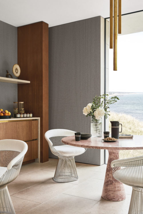 Un charmant coin petit-déjeuner dans un hôtel de bord de mer, avec une grande fenêtre offrant une vue pittoresque sur les dunes et la mer. Une table ronde en marbre occupe le devant de la scène, entourée de fauteuils beiges accueillants. La table est surmontée d'un vase en verre contenant un bouquet de fleurs, éclairé par un plafonnier doré à trois tubes. Les murs sont élégamment ornés de revêtements muraux Rivage d'Omexco en gris foncé, présentant un design uni subtil qui renforce l'atmosphère sereine de la retraite côtière. Ces revêtements muraux sont non seulement exempts de PVC et nettoyables à l'eau de Javel, mais ils sont également plus performants en ce qui concerne la facilité de nettoyage.