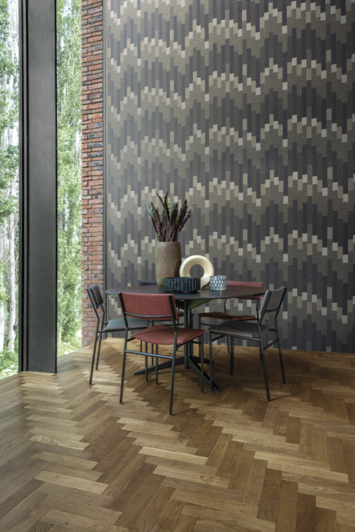 Salle à manger avec une mosaïque de fines paillettes de mica sur le mur. Les formes forment un motif géométrique rythmé et saisissant dans des tons de violet foncé, de gris et de beige. Il s'agit du modèle "Cadence" de la collection Moonstone d'Omexco. Nous voyons une table de salle à manger avec 4 chaises, quelques décorations sur la table, une grande fenêtre sur la gauche et un sol en bois à chevrons.