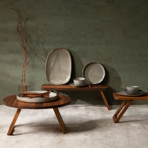 Image décorative d'une table basse ronde et d'une table basse rectangulaire en bois posées sur un grand tapis blanc cassé. Des assiettes en céramique de différentes formes et tailles sont posées sur les tables basses et, à gauche, un vase en verre est posé sur le sol et contient des brindilles décoratives. À l'arrière-plan, nous voyons la collection de revêtements muraux en liège "Casca" d'Omexco, et plus particulièrement le motif "tactility" dans une couleur vert mousse. Il s'agit d'un faux uni, résultat d'un patchwork d'éléments rectangulaires en liège qui crée une variété de teintes différentes sur le mur.