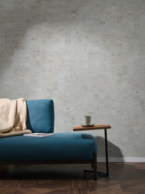 Image de décoration d'un canapé bleu avec un plaid beige accroché au dossier. À droite, une table d'appoint minimaliste sur laquelle est posée une tasse en céramique. En arrière-plan, nous voyons la collection de revêtements muraux en liège "Casca" d'Omexco, plus particulièrement le motif "tactility" dans une couleur bleu-gris-vert. Il s'agit d'un revêtement mural faussement uni, résultat d'un patchwork d'éléments rectangulaires en liège qui crée une variété de teintes différentes sur le mur.