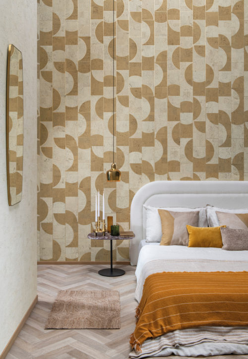 Image d'une chambre à coucher élégante avec un sol clair à chevrons, un lit blanc avec des oreillers et une couverture ocre, un miroir et une lampe à suspension moderne dorée. Une table d'appoint en marbre noir offre un contraste élégant avec les couleurs claires, tandis qu'un revêtement mural géométrique blanc et jaune ajoute une touche ludique. La mosaïque de formes géométriques contrastées est en liège et fait partie de la collection de revêtements muraux "Casca" d'Omexco. L'effet global est un espace frais et moderne qui est à la fois accueillant et visuellement intriguant.