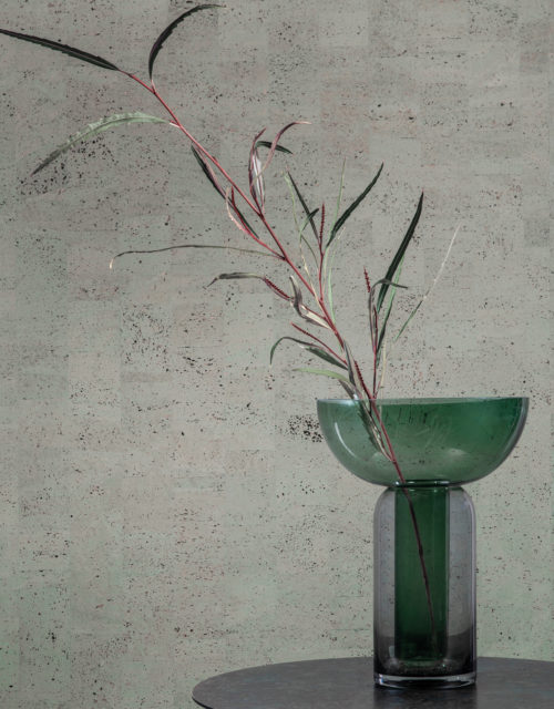 Gros plan sur un vase en verre transparent vert contenant une tige de plante, posé sur une table d'appoint ronde noire. En arrière-plan, nous voyons la collection de revêtements muraux en liège "Casca" d'Omexco, et plus particulièrement le motif "tactility" dans une couleur gris-vert bleuté. Il s'agit d'un revêtement mural faussement uni, résultat d'un patchwork d'éléments rectangulaires en liège qui crée une variété de teintes différentes sur le mur.