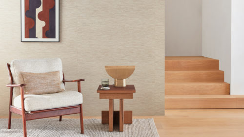 Dans cette image, un fauteuil vintage avec un coussin beige est placé sur un tapis beige à côté d'une table d'appoint en bois avec une lampe de table dorée et un verre d'eau. Sur la droite, un escalier en bois de 4 marches. Un tableau de formes géométriques dans les tons marine, blanc et rouille est accroché au mur sur lequel on peut voir les revêtements muraux Loom Stories d'Omexco. Cet élégant tissage de lin et de coton est présenté ici dans des tons apaisants de beige et de marron clair. La combinaison d'éléments vintage et modernes ajoute une touche d'élégance et de sophistication à ce coin douillet.