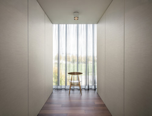 Couloir avec chaise centrale en bois devant une fenêtre. Deux lampes rondes blanches au plafond. Revêtements muraux de type portfolio sur panneaux sur les murs.