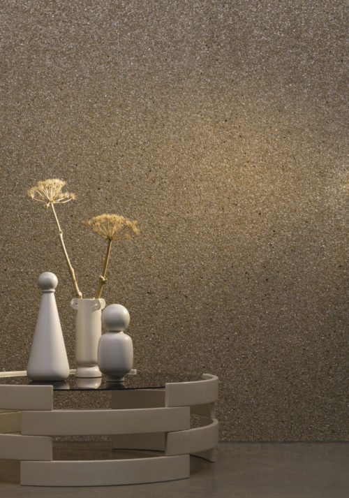 Petit décor avec des revêtements muraux Omexco unis. Le revêtement mural est composé de fines paillettes de mica opalescentes, dans des tons de brun doré. Une toile de fond parfaite pour la table basse blanche avec des vases blancs en céramique et deux fleurs séchées.