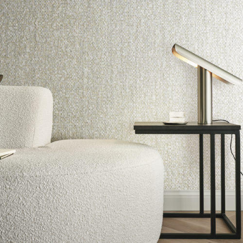 Gros plan sur un salon confortable, avec un canapé en tissu bouclé beige orné d'un coussin marron. Le parquet à chevrons complète les plinthes classiques, créant une esthétique intemporelle. Une table d'appoint noire se trouve à proximité, ornée d'une lampe de table dorée minimaliste. En toile de fond, les revêtements muraux en nacre capiz de la collection Archipelago d'Omexco présentent des nuances chatoyantes de beige, ajoutant une touche d'élégance et de sophistication à cet espace intime.