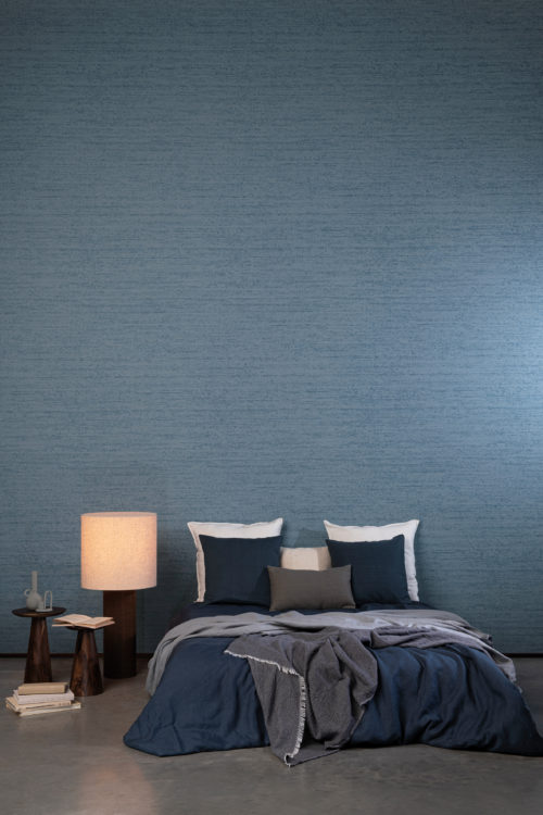 Chambre à coucher moderne et impressionnante avec un sol en béton, des oreillers et des draps bleu marine et des tables de chevet en bois de forme conique. Sur les murs de cet espace contemporain, nous voyons le motif "Timber" de la collection de revêtements muraux High Performance Patina d'Omexco. Ce motif très texturé s'inspire de la structure du bois brûlé, un aspect très populaire de nos jours. Le revêtement mural bleu denim est posé horizontalement.