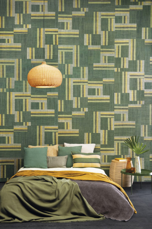 L'artisanat authentique incarne l'audacieuse collection Atelier conçue par les esprits créatifs d'Omexco. Cette image montre un impressionnant patchwork vert de raphia tissé à la main. Le mur parfait dans cette chambre à coucher tropicale avec des accents de couleur ocre, une table de chevet en métal vert, une feuille de plante exotique et des coussins de lit douillets.