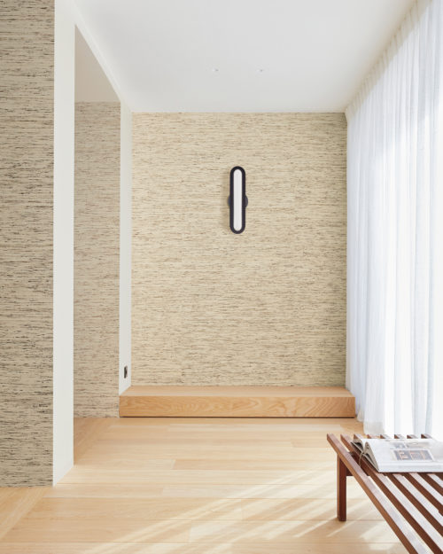 Un couloir accueillant dans une maison moderne, avec des rideaux blancs et transparents qui permettent à la lumière naturelle d'inonder l'espace. Le parquet ajoute de la chaleur et de la texture à l'espace, tandis qu'un élégant luminaire mural apporte une touche de style contemporain. Sur la droite, un banc en bois permet de s'asseoir confortablement et le papier peint texturé crée un contraste visuel intéressant. Ici, nous voyons les revêtements muraux Loom Stories d'Omexco dans des tons de beige et de marron clair, avec un motif subtil qui ajoute de la texture au mur. L'esthétique sauvage est créée par l'irrégularité des fils soyeux.