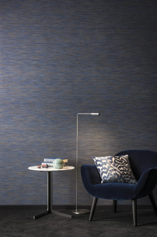 Fauteuil bleu nuit en velours, confortablement installé devant le revêtement mural Omexco Portfolio, également bleu. Liseuse, table d'appoint avec des livres et une figue.