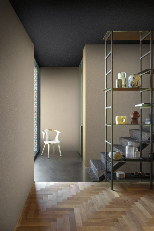Intérieur aux couleurs neutres. Sol en bois à chevrons. La grande fenêtre et la chaise blanche au fond apportent de la luminosité à l'espace. Les murs sont recouverts de revêtements muraux Moonstone d'Omexco, fabriqués à partir de mica naturel. Le motif est simple, mais chatoyant, car le mica est une pierre semi-précieuse à l'éclat séduisant.