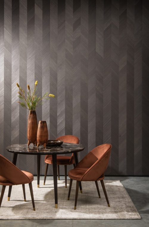 La collection Sycamore d'Omexco : un glamour doux qui reflète notre désir d'une connexion plus profonde avec la nature. Sur cette photo, l'élégance intemporelle du motif à chevrons est associée à une table en marbre brun, à des chaises et à des vases de couleur rouille.