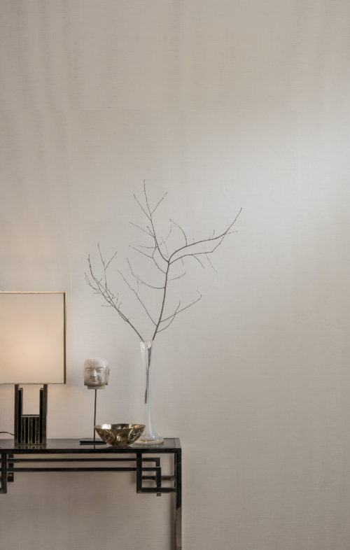 Table art déco en laiton présentant une statue de tête, une fine branche et une lampe blanche. Le revêtement mural en lin Shades of Pale vient parfaire l'ensemble.