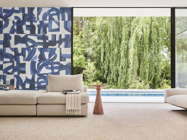 Un salon époustouflant où l'on retrouve le revêtement mural Rivage d'Omexco dans une teinte bleue captivante avec des coups de pinceau artistiques. Le design tisse habilement des échos de cordes et de nœuds maritimes, rendant ainsi hommage à la complexité de la navigation. Au premier plan, un gros plan sur un canapé beige et une table d'appoint minimaliste en bois exsudent l'élégance moderne. Au-delà, une fenêtre ouverte révèle une vue rafraîchissante sur une piscine, ajoutant à l'impression générale de tranquillité et de sophistication de cet espace magnifiquement décoré.