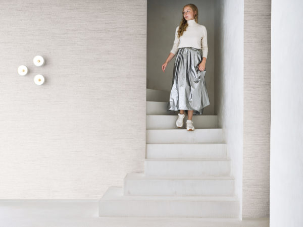 Un intérieur minimaliste et contemporain mettant en avant la collection Pure d’Omexco, référence PU251, nommée Delicate. Ce papier peint raffiné présente un raffia fin sublimé par des fils métalliques subtils, combinant une texture naturelle avec une touche d’éclat pour une esthétique sophistiquée. Une femme descend un escalier blanc vêtue d’un pull à col roulé crème et d’une jupe métallisée argentée, apportant une élégance moderne à cet espace serein. L’arrière-plan accentue l’ambiance paisible avec sa texture discrète mais complexe, idéal pour les intérieurs minimalistes valorisant le luxe subtil et l’artisanat.