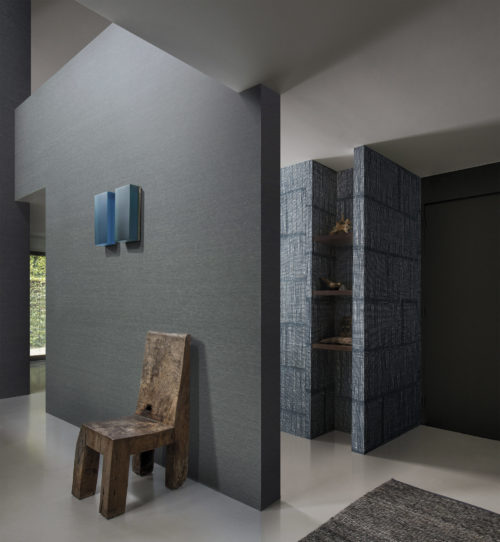 Couloir avec une chaise en bois robuste. Au-dessus, une œuvre d'art en verre bleu. Un revêtement mural en raphia bleu forme une bibliothèque.