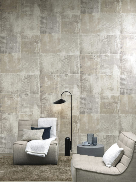 Sur cette image, nous voyons un mur d'accent avec des carrés organiquement flétris imprimés sur une texture de fines paillettes de mica opalescentes. Ces revêtements muraux rappellent les graffitis délavés par le temps. Le motif s'appelle "Mémoires" et appartient à la collection Moonstone d'Omexco. Deux canapés à une place sont placés devant le mur et le sol est recouvert d'une moquette grise en peluche. Les tons de cette pièce sont neutres : beige, gris clair et gris bleu.
