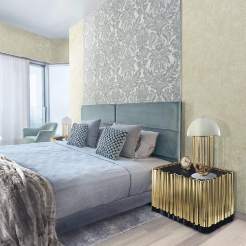 Chambre à coucher bleu clair avec des tableaux texturés et un plaid en fourrure. Table de nuit dorée avec lampe dorée. Au mur, un revêtement mural damassé bleu clair d'Omexco.