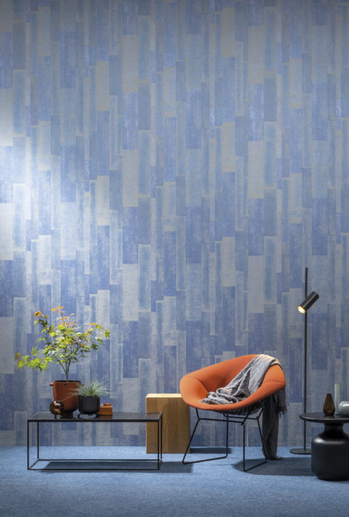 Impressionnant motif géométrique en bleu électrique de la collection Omexco Loft. Le revêtement mural est posé verticalement et associé à une chaise longue orange et à une plante d'érable japonaise.