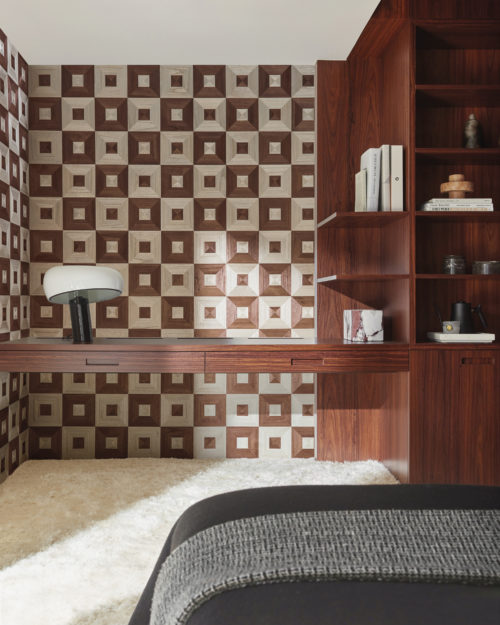 Une chambre à coucher sereine avec un tapis blanc en peluche et des draps gris foncé sur le lit. Une bibliothèque en bois foncé ornée d'une lampe de table élégante et de livres se trouve contre le mur. La lumière du jour éclaire la pièce. Le mur est orné de la collection de marqueterie Imperialis d'Omexco, qui présente un motif de marqueterie en damier alternant des tons contrastés de blanc et de bordeaux, créant un effet graphique saisissant.