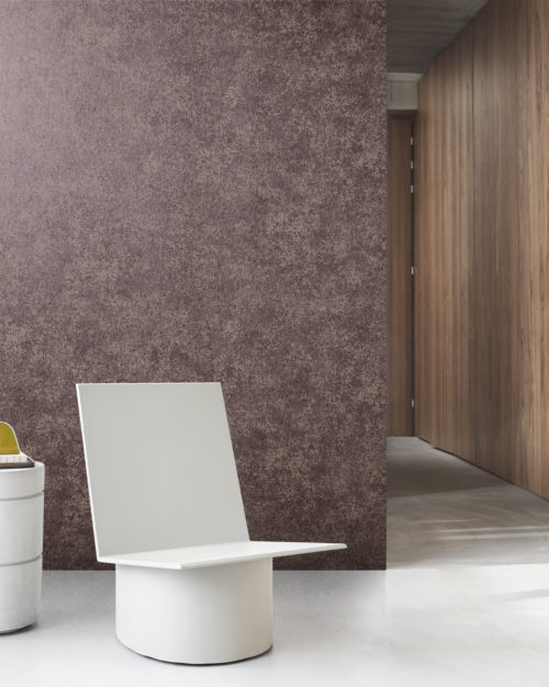 Une pièce minimaliste avec un revêtement mural intissé imprimé effet patine de la collection "Edition" d'Omexco. Le mur a une apparence texturée dans des tons de violet et de marron. Au premier plan, il y a une chaise blanche moderne au design géométrique. À gauche, un conteneur cylindrique blanc est partiellement visible. À l'arrière-plan, un couloir avec des murs en bois s'étend au loin.
