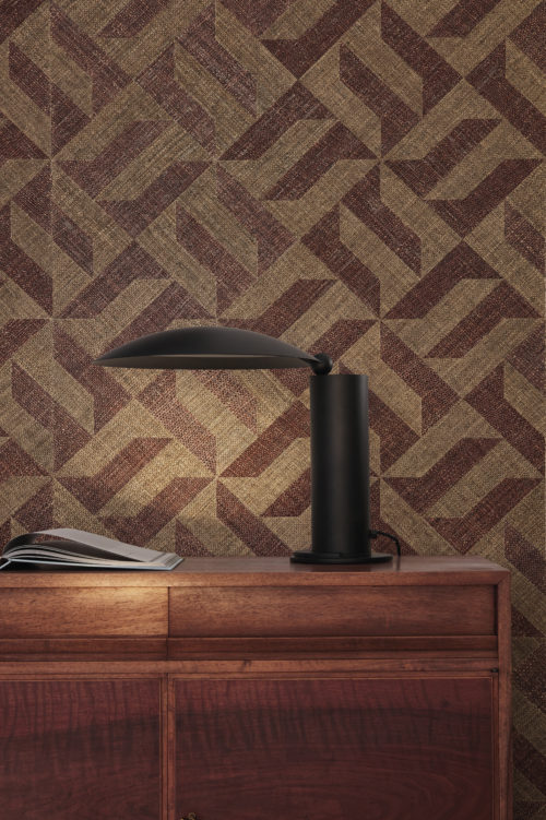 Gros plan sur une armoire en bois surmontée d'une élégante lampe de table noire, accompagnée d'un livre ouvert. La toile de fond présente les revêtements muraux Intuition d'Omexco - un patchwork de raphia avec un motif géométrique captivant aux formes bordeaux et marron clair. La composition respire la sophistication et la chaleur, créant une ambiance à la fois confortable et élégante