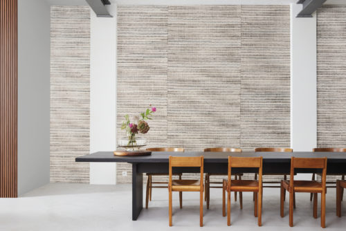 Une salle à manger chic avec un sol en fonte grise, comprenant une longue table noire entourée de huit chaises en bois. Les hauts murs et le plafond métallique de style industriel ajoutent une ambiance de loft. Un vase contenant des fleurs éclatantes sert de centre de table. La collection de revêtements muraux Natural "Intuition" d'Omexco est exposée en toile de fond : un tissage de bakbak, de bacnoc et de fils de papier avec des nuances de beige et de brun, qui renforce l'attrait contemporain et élégant de la pièce.