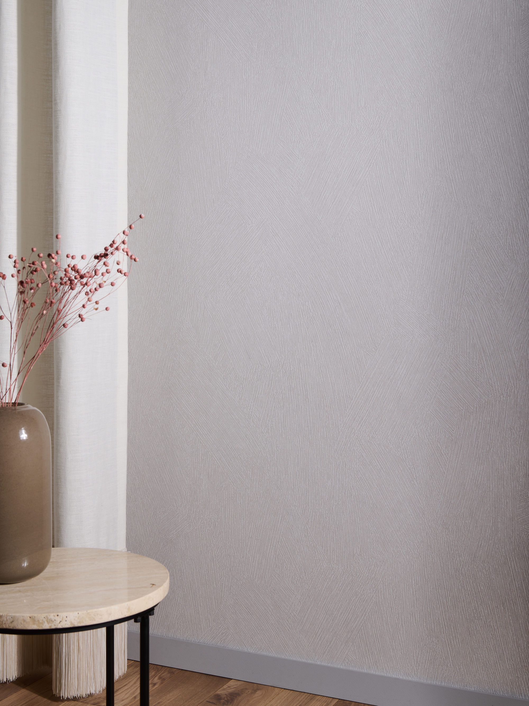 Image d'un coin de salon aéré et contemporain avec une table ronde en bois et un vase en céramique marron clair rempli de fleurs roses séchées. On voit un rideau en lin blanc sur la gauche. Un parquet en bois crée une base naturelle tandis qu'un revêtement mural Omexco neutre à effet craie brossée sert de toile de fond. Ces revêtements muraux High Performance Patina répondent aux normes techniques essentielles tout en étant respectueux de l'environnement.