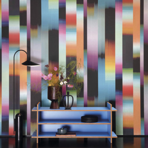 Décoration d'intérieur funky inspirée des années soixante-dix. Faites entrer dans votre maison l'ambiance unique d'une période incroyable avec le design du revêtement mural "Time-lapse" de la collection "Atelier" d'Omexco. Cette interprétation moderne d'un tissage Ikat présente des rayures dynamiques dans un dégradé de couleurs orange, turquoise, rose, violet, noir et rouge. La toile de fond parfaite pour une armoire ouverte bleu électrique avec des étagères et un lampadaire noir minimal.