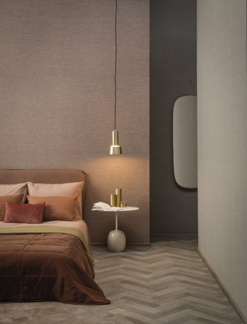 Sur les murs de cette chambre d'hôtel : le motif Abaca dans les tons blush et beige de la collection High Performance Textures d'Omexco. Il s'agit d'un revêtement mural intissé semi-uni à la texture intrigante, présenté à l'horizontale. Le revêtement mural est associé à un lit douillet, une lampe pendulaire, des oreillers orange et beige, un miroir et une table d'appoint.