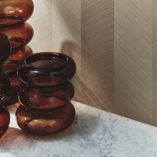 Gros plan sur une luxueuse surface en marbre sur laquelle sont disposés trois vases bruns en verre. À l'arrière-plan, la collection de revêtements muraux en marqueterie Imperialis d'Omexco constitue une toile de fond étonnante, mettant en valeur la beauté subtile d'un placage de bois fin dans un motif à chevrons, incrusté à la main de manière experte. La lumière du jour éclaire le mur d'une douce lumière, rehaussant l'élégance de la scène.