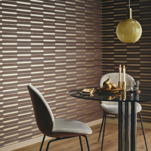 Un coin de salle à manger confortable avec un sol en parquet et une table ronde noire flanquée de deux chaises grises confortables. Au-dessus de la table, une lampe ronde en verre jaune éclaire l'espace. Au mur, la collection de revêtements muraux en placage de bois Imperialis d'Omexco crée une toile de fond saisissante avec son motif géométrique de bois de bambou brun foncé sur une feuille d'or gaufrée.