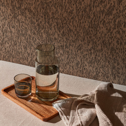 Gros plan sur un petit plateau en bois contenant une cruche transparente et un verre, tous deux remplis d'eau, ainsi qu'un torchon de cuisine beige. Les objets sont placés sur un lit de jour moelleux, également de couleur beige. Sur le mur de cette pièce sereine aux tons neutres, nous voyons un revêtement mural texturé avec un motif organique marron. Ces revêtements muraux naturels ont l'apparence du cuir, mais sont en fait fabriqués à partir d'écorce de chêne-liège. Ils appartiennent à l'exquise collection "Casca", fabriquée à la main par Omexco. Le jeu intéressant de la lumière naturelle et de l'ombre crée une atmosphère calme et accueillante.