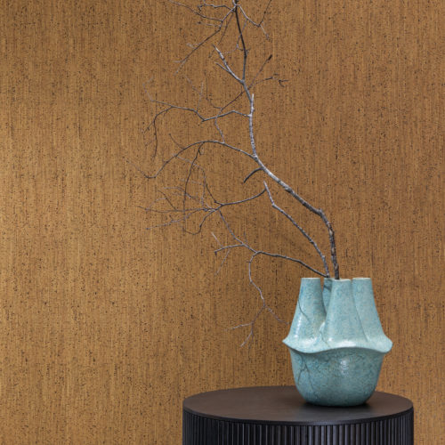 Gros plan sur un vase en céramique turquoise tenant une petite branche d'arbre, posé sur une table d'appoint ronde en bois noir. En arrière-plan, nous voyons la collection de revêtements muraux en liège "Casca" d'Omexco, et plus particulièrement le motif "enhance" dans une teinte brun caramel. Ce revêtement mural naturel, fabriqué à la main, est le résultat de plusieurs fines couches de liège empilées et pressées. Elles sont ensuite découpées en feuilles très fines. Le résultat visuel est comme si les rayures étaient imprimées.
