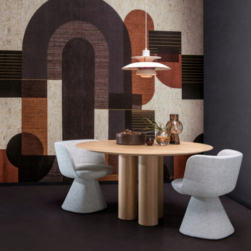 Exemple d'une salle à manger avec un revêtement mural au graphisme audacieux présentant un motif de formes géométriques dans des tons gris, noir, orange brûlé et blanc. Ce motif panoramique est imprimé numériquement sur du liège et fait partie de la collection de revêtements muraux "Casca" d'Omexco. Au centre de la pièce se trouve une table à manger ronde en bois au-dessus de laquelle est suspendue une lampe blanche. Des éléments décoratifs tels que de petits vases en verre et un bol rempli de noix sont placés sur le plateau de la table. Deux chaises de salle à manger d'un gris doux sont disposées autour de la table. L'esthétique générale est moderne avec une touche de chaleur et de texture.