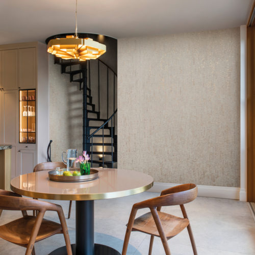 Exemple de cuisine lumineuse et aérée avec de grands carreaux gris clair, des armoires de cuisine blanches, une table à manger ronde avec trois chaises en bois et un escalier en colimaçon noir en arrière-plan. Une lampe suspendue dorée ajoute une touche d'élégance et de chaleur à l'espace, tandis qu'un revêtement mural beige métallisé ajoute une texture subtile et un intérêt visuel. Ici, nous voyons la collection de revêtements muraux en liège "Casca" d'Omexco. L'effet global est une cuisine moderne et accueillante, parfaite pour la préparation des repas et les dîners décontractés.