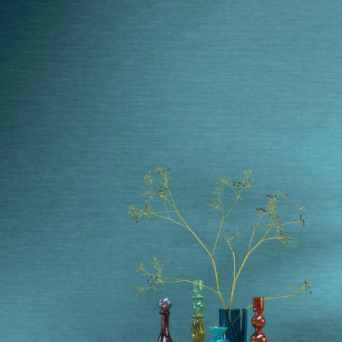 Gros plan sur un mur turquoise vif. Il s'agit de l'uni le plus populaire d'Omexco, imprimé sur un intissé texturé pour lui donner un aspect textile familier. Le motif de ce revêtement mural s'appelle "Silky" et appartient à la collection tropicale "Birds of Paradise". Devant le mur, nous voyons une table d'appoint dorée sur laquelle sont posés des vases en verre colorés de différentes hauteurs et formes.