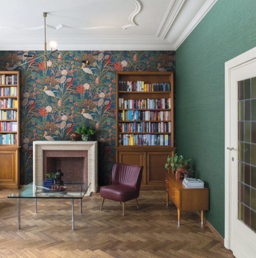 Salon de style parisien avec un haut plafond, une cheminée et un sol à chevrons en bois foncé. Derrière la grande bibliothèque se trouve un revêtement mural au motif botanique composé de feuillages, de fleurs et d'échassiers. Sur un fond bleu foncé, on voit ici l'ibis rouge et la spatule blanche. Il s'agit des revêtements muraux tropicaux "Birds of Paradise" d'Omexco.