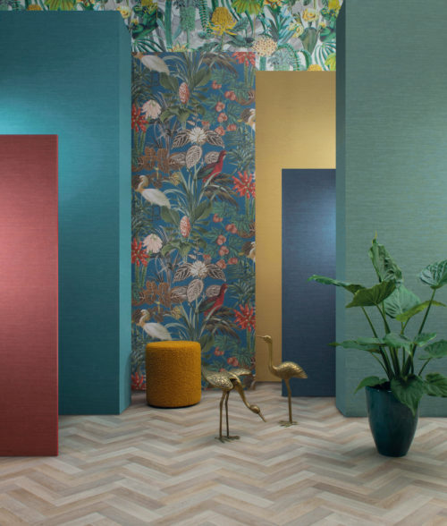 Espace intéressant avec un sol clair à chevrons et six panneaux colorés de différentes hauteurs, allant de la mi-hauteur à la hauteur du plafond. Au sol, on trouve un pouf rond ocre, une grande plante d'intérieur tropicale dans un pot en céramique vert et deux statues dorées de grues. Les panneaux sont décorés de revêtements muraux unis de couleur jaune, turquoise et bleu foncé, ainsi que de motifs tropicaux représentant de fabuleuses fleurs, des feuilles luxuriantes et de magnifiques oiseaux. La collection de revêtements muraux "Birds of Paradise" d'Omexco est illustrée ici.