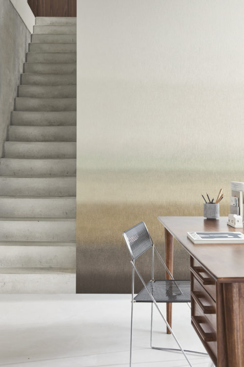 Photo décor avec un escalier en béton minimaliste sur le côté gauche de l’image. Le mur de fond présente la collection intissé imprimé « Edition » d’Omexco, un design intemporel et doux en dégradé passant du beige, vert clair et marron clair au marron foncé. À droite, il y a un bureau en bois moderne avec quelques objets, dont un magazine, une tasse avec des crayons et un petit objet décoratif. Une chaise en métal élégante est placée devant le bureau. Le sol est de couleur claire, complétant l’esthétique moderne et minimaliste de l’espace.