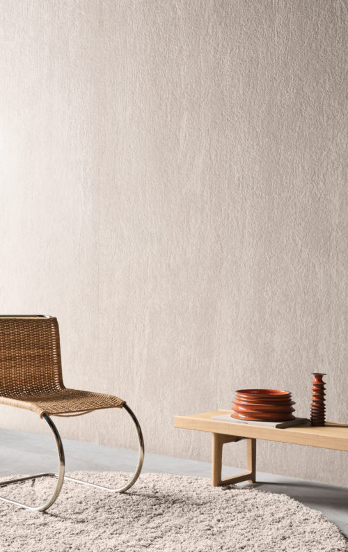 Un intérieur accueillant mettant en avant la collection Pure d’Omexco, référence PU401, composée de velours beige à poils longs. Ce revêtement mural offre une texture luxueusement douce et moelleuse qui dégage chaleur et sophistication. La scène met en valeur une chaise contemporaine en osier avec des pieds chromés, posée sur un tapis texturé et doux. À droite, un banc en bois minimaliste accueille des bols en terre cuite et un vase sculptural, associant des matériaux naturels à des tons terreux. Ce design élémentaire capture la beauté tactile du velours, sublimant l’ambiance sereine et raffinée.