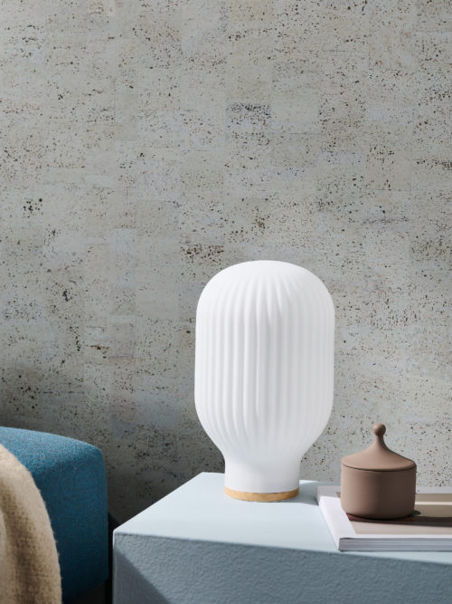 Gros plan sur la lampe de table Baja d'Habitat. La lampe blanche, en forme de montgolfière, est posée sur une table d'appoint bleu clair. Sur la gauche, nous voyons le coin d'un canapé bleu avec un plaid beige. En arrière-plan, la collection de revêtements muraux en liège "Casca" d'Omexco est présentée, plus particulièrement le motif "tactility" dans une couleur gris-vert bleuâtre. Il s'agit d'un revêtement mural faussement uni, résultat d'un patchwork d'éléments rectangulaires en liège qui crée une variété de teintes différentes sur le mur.