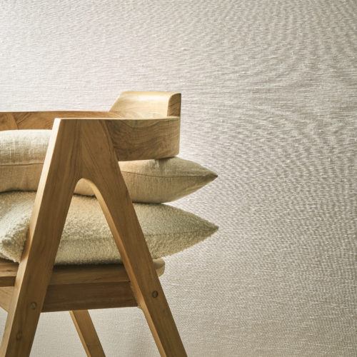 Une composition sereine mettant en avant la collection Pure d’Omexco, référence PU362, composée de fils textiles très fins, fabriqués à partir de matériaux nobles tels que le lin. Ce revêtement mural dégage une sophistication subtile grâce à sa texture raffinée, ajoutant une élégance discrète à l’espace. La scène présente une chaise en bois aux lignes angulaires épurées, surmontée de coussins empilés dans des tons naturels et une variété de textures, y compris du lin doux et du tissu bouclé. L’arrière-plan neutre et les matériaux naturels s’harmonisent pour créer une atmosphère chaleureuse et accueillante, parfaitement adaptée aux intérieurs modernes et minimaliste.